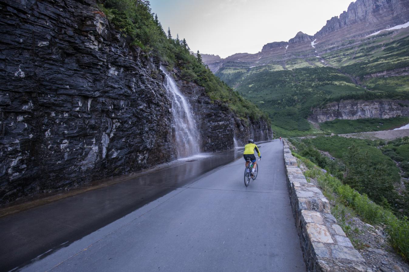 man on bike on road in Montana with waterfall and mountains in background